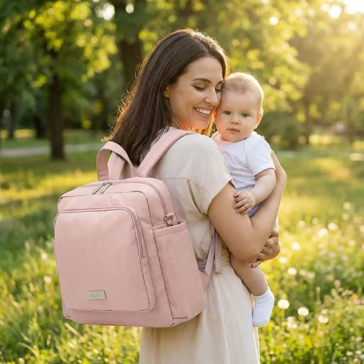 Maman en campagne avec bébé dans les bras et Sac à dos à langer rose sur les épaules